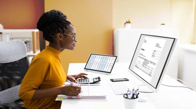 A professional woman with short dark hair and glasses, wearing a bright yellow top, sits at a modern desk working on financial documents. She is using a desktop computer with spreadsheets visible on the screen while also referencing printed accounting documents. The clean, well-lit workspace includes office supplies and represents a contemporary accounting professional in action