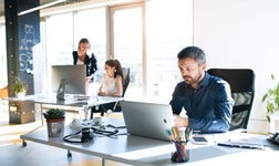 A professional man working on a laptop in a sunlit modern office with colleagues in the background.