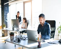 A professional man working on a laptop in a sunlit modern office with colleagues in the background.