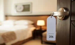 Open hotel room door with room 206 key in door on foreground and double bed and side table out of focus in background