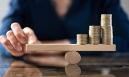 A person's fingers carefully adjusting a wooden balance scale with ascending stacks of coins, depicting financial stability and investment growth.