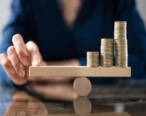 A person's fingers carefully adjusting a wooden balance scale with ascending stacks of coins, depicting financial stability and investment growth.