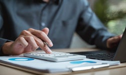 Man sitting at desk with files, calculator, and laptop.