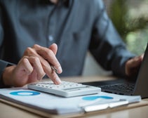 Man sitting at desk with files, calculator, and laptop.