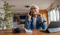 Middle aged woman sitting at dining room table in front of iPad and coffee cup laughing while talking on the phone.
