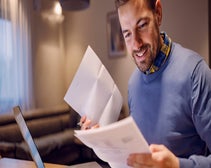man smiling and holding invoices printed on paper while sitting at desk in front of laptop