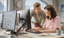 two people in a modern office looking at a monitor screen, while another monitor behind has CRM software displayed
