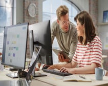 two people in a modern office looking at a monitor screen, while another monitor behind has CRM software displayed