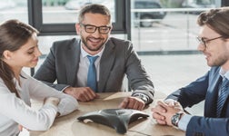 Three business people sitting at desk talking on conference phone.