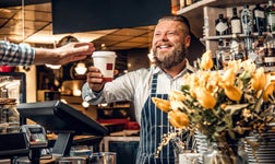 Smiling male barista hands coffee in takeaway cup to customer at counter