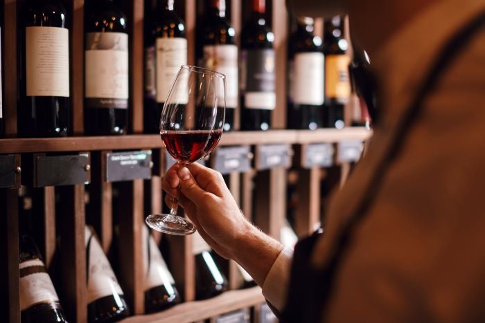 Caviste tenant un verre de vin rouge devant des étagères remplies de bouteille.