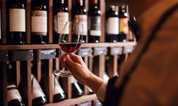 Caviste tenant un verre de vin rouge devant des étagères remplies de bouteille.