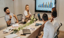 five people around a long table, with documents and laptops sitting in front of each person and a screen in the background showing a chart