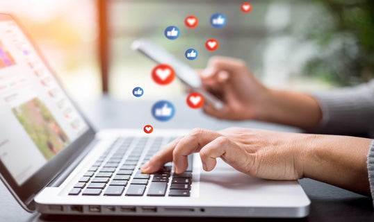 Hands typing on a laptop keyboard with floating social media interaction icons, depicting active engagement on social media platforms.