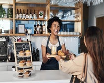 Smiling female barista serving female customer croissant and cofee accross the counter at a cafe/coffee shop