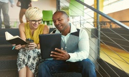 Smiling woman holding a notebook looks at an ipad screen with a man