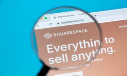 Magnifying glass held over laptop showing the Squarespace website builder homepage promoting online sales capabilities.