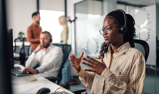 person on the phone using a headset for VoIP calling in a dynamic office setting, three people behind blurry