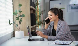 Business woman is using a tablet and talking on the phone while sitting at her desk in a modern office