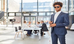 businessman wearing a suit and dark shades holds a cane standing in an office setting