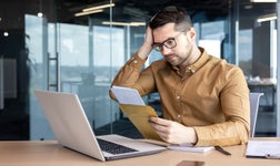 man holding a letter looks at a laptop in an office seems stressed