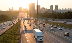 cars, trucks and vans on US and Canadian highway in front of city skyline bathed in sunlight