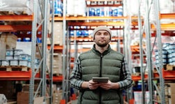man holding an iPad stands in a warehouse full of assets on shelves that need tracking