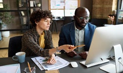 Businesswoman pointing at computer monitor and discussing presentation together with her colleague at at office