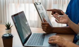 person using laptop on a wooden table alongside another person holding a clipboard with graphics on it