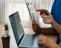person using laptop on a wooden table alongside another person holding a clipboard with graphics on it