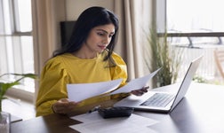 woman doing accounting paperwork at home, sitting at table with calculator and laptop, reviewing documents.
