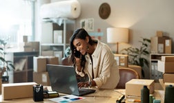 Smiling business woman on phone checking laptop in home office surrounded by mail boxes.