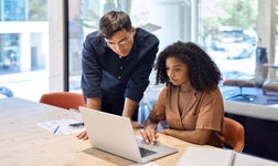 Woman sitting at desk and man standing over her both looking at laptop screen.