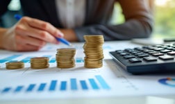 Business person review financial data. Stacks of coins represent budget growth. Charts, graphs show financial performance. Calculator sits nearby for budget calculations. Person reviews investment