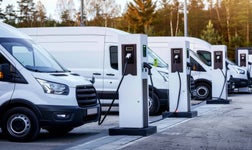 A row of electric vans are plugged in and charging at a public charging station.